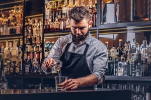 stylish brutal barman in a shirt and apron makes a cocktail at bar counter background